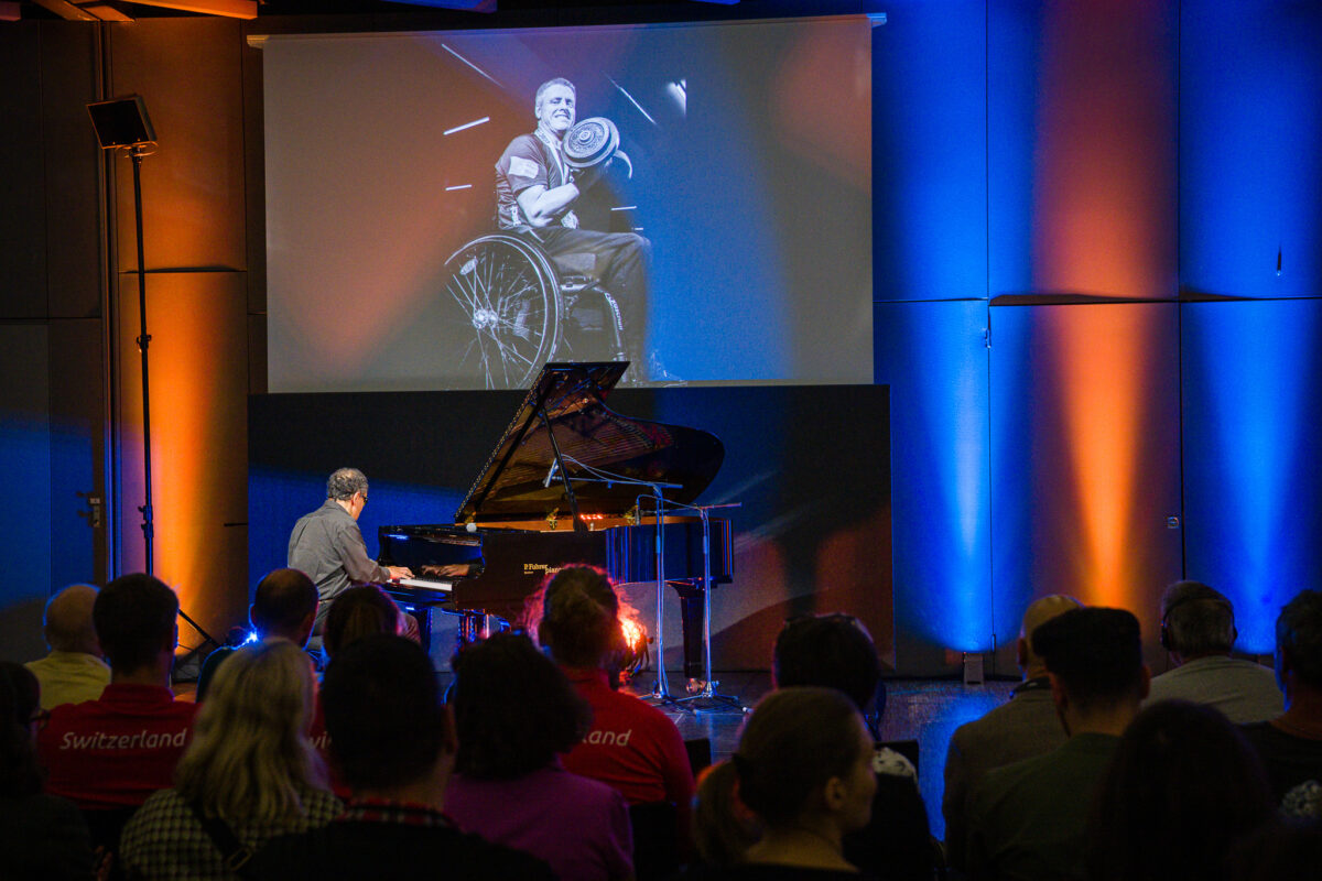 Des gens de dos regardent et écoutent Moncef Genoud jouer au piano. Sur un écran géant, on voit une image en noir et blanc d'homme en fauteuil roulant qui fait des haltères.