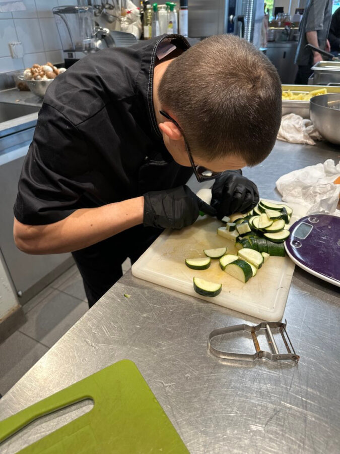 Une personne tête baissée coupe des courgettes dans une cuisine professionnelle