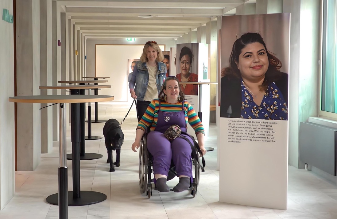 Une femme en chaise roulante parcourt une exposition de photographie, une autre femme marche derrière elle avec un chien en laisse