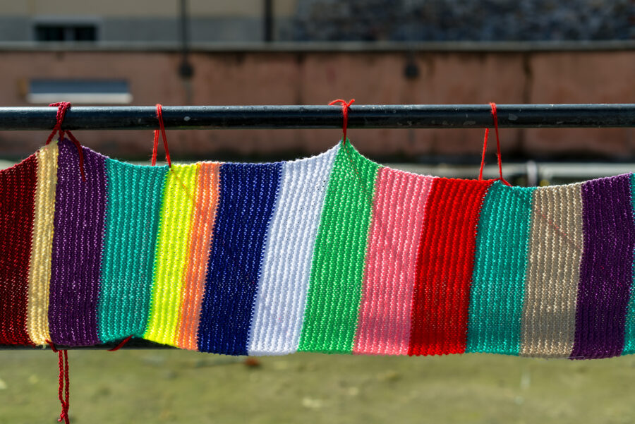 Une création en laine multicolore suspendue à une barrière en extérieur et qui fait penser à un bel arc-en-ciel.