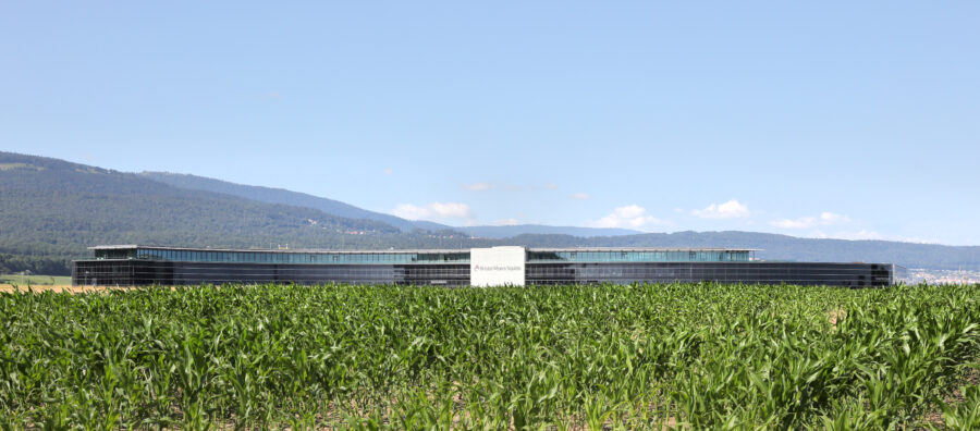 Photo en format paysage du bâtiment de BMS. Le bâtiment est situé au loin, entre un parterre d'herbe et le ciel bleu. On aperçoit le début de la montagne de Boudry en haut à gauche.