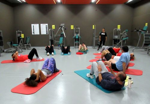 Une salle avec plusieurs engins de musculation. Plusieurs personnes forment un cercle et sont engagées dans des activités physiques sur des tapis d'exercice. Un coach est debout et donne des instructions