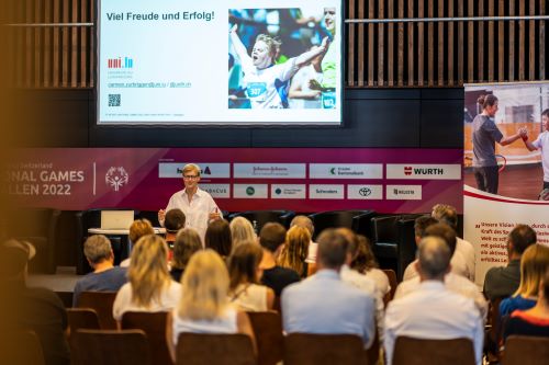 Une salle de conférence avec un conférencier qui fait face. Derrière lui un écran projette une présentation avec une photo d'un athlète qui lève les bras à l'arrivée d'une course. Les spectateurs sont assis sur des chaises brunes, de dos.