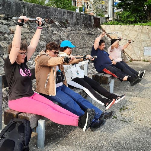 4 personnes sont assises sur des bancs, les jambes tendues à l'horizontal et tenant des bâtons de Nordic Walking au-dessus de leur tête. Elles sont en train d'effectuer des mouvements d'échauffement.