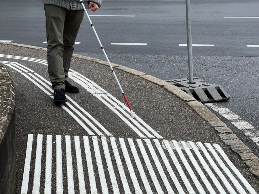 Photo des jambes d'une personne en marche avec une canne blanche qui suit les lignes de guidage sur un trottoir.