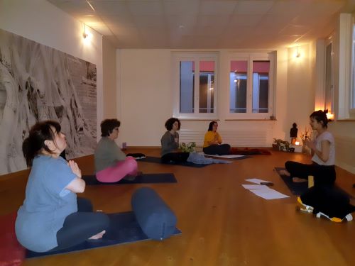 Un groupe de personnes assises sur des tapis d'exercice. Elles participent à une séance de pilates. Une personne guide la session au centre tandis que d'autres sont orientées vers elle. Des coussins sont déposés à l'avant des tapis.