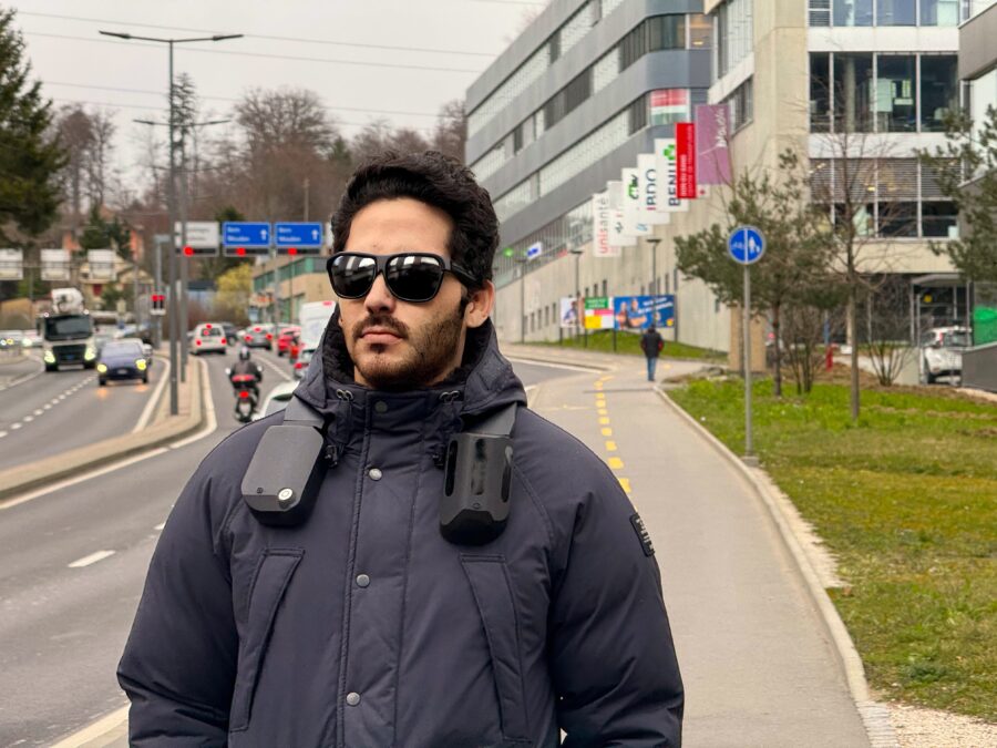 Un utilisateur, Amine, porte un harnais biped sur les épaules. Il marche sur un trottoir, avec le trafic qui défile derrière lui, suggérant un environnement urbain que l’appareil aide à déchiffrer.