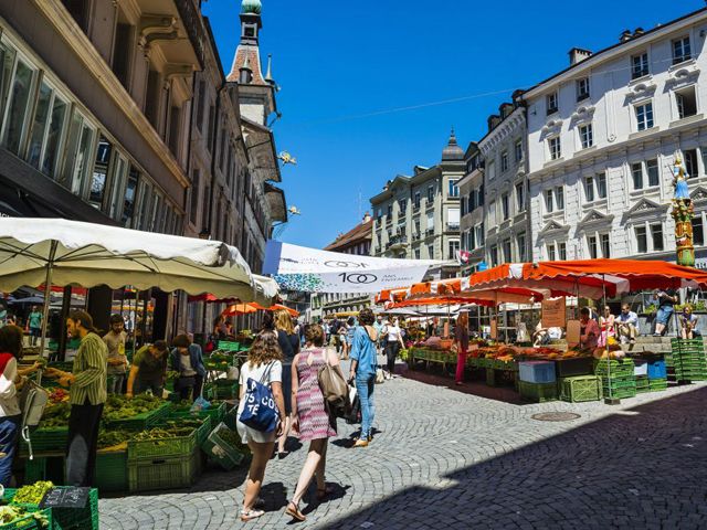Photo montrant la Place de la Palud à Lausanne, avec des stands de marché et des gens qui circulent au marché.