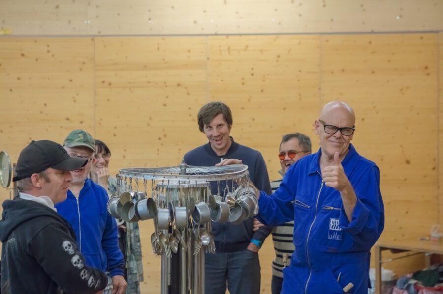 C’est une photo avec des personnes qui sont debout et qui sourient. Elles sont autour d’un instrument de musique fabriqué avec une roue de vélo, des boites de conserve et des fourchettes en métal.